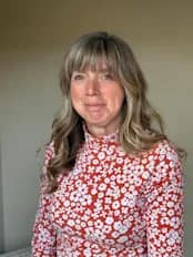 Paula Fowle has long light brown hair and is wearing a white dress with brown flowers