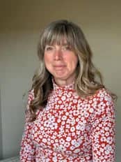 Paula Fowle has long light brown hair and is wearing a white dress with brown flowers