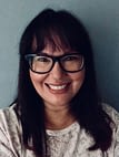 Senior Associate Mish Seabrook has long dark hair and is wearing black glasses and a white and grey top